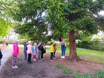 Dzieci w ogrodzie, stoją pod drzewem, a bibliotekarka pokazuje im domek  na owady