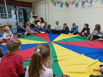 Dzieci podczas zajęć siedzą w kole na kolorowej chuście i słuchają bibliotekarki, czytającej książkę