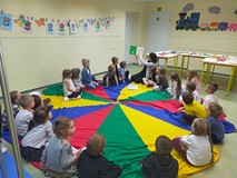 Dzieci siedzą na kolorowej chuście animacyjnej i słuchają opowiadania
