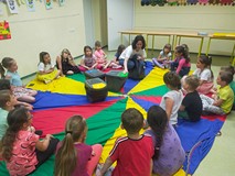 Dzieci siedzą na kolorowej chuście animacyjnej, a bibliotekarka pokazuje pojemniki na szkło, papier 