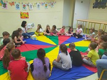 Dzieci siedzą na kolorowej chuście animacyjnej i słuchają opowiadania