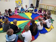 Dzieci siedzą na chuście i słuchają książeczki czytanej przez bibliotekarkę