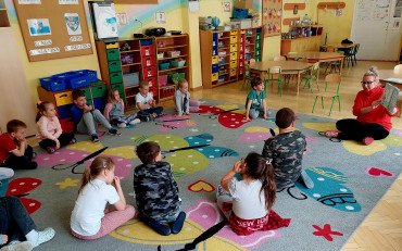 Dzieci siedzą na podłodze i słuchają czytającej książeczkę pt. 