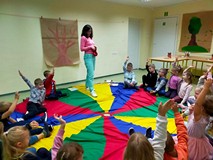Dzieci siedzą na kolorowej chuście animacyjnej, a bibliotekarka pokazuje woreczek z zagadkami