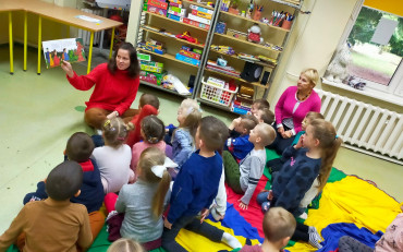 Dzieci podczas zajęć siedzą w kole na kolorowej chuście i słuchają opowiadania,...