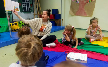 Dzieci podczas zajęć na kolorowej chuście i słuchają opowiadania, czytanego przez...