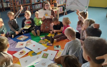 Dzieci siedzą na chuście animacyjnej i słuchają czytanego przez bibliotekarkę opowiadania