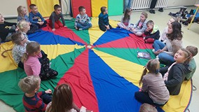 Dzieci siedzą na kolorowej chuście animacyjnej i rozmawiają z bibliotekarką