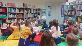 Dzieci siedzą na kolorowej chuście animacyjnej, a bibliotekarka czyta opowiadanie