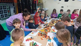 Dzieci stoją przy stolikach i wykonują prace plastyczną