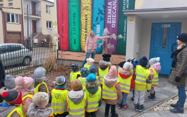 Dzieci stoją przd biblioteką słuchając bibliotekarki