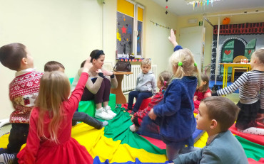 Dzieci podczas zajęć siedzą w kole na kolorowej chuście i rozmawiają z bibliotekarką