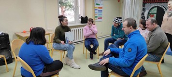 Uczestnicy zajęć siedzą na krzesełkach i słuchają opowiadania czytanego przez bibliotekarkę