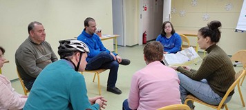 Uczestnicy zajęć siedzą na krzesełkach i słuchają opowiadania czytanego przez bibliotekarkę