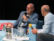 Na scenie siedzą Grzegorz Śliżewski i prowadzący spotkanie Piotr Polechoński