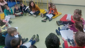Dzieci podczas zajęć siedzą w kole na kolorowych materacykach i prezentują wykonane prace plastyczne