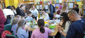 dzieci z rodzicami siedzą przy stole i ozdabiają wielkanocne jaja pod nadzorem bibliotekarki