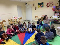 Dzieci siedzą na kolorowej chuście, a bibliotekarka czyta książkę