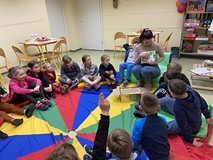 ieci podczas zajęć siedzą w kole na kolorowej chuście, a bibliotekarka pokazuje ilustrację