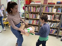 Bibliotekarka trzyma w rękach karton, chłopiec stojący obok bibliotekarki wrzuca do niego białą kope