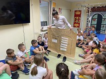 Bibliotekarka pokazuje dzieciom plakat. Dzieci siedzą na kolorowych materacykach