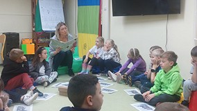 Dzieci siedzą na materacykach i słuchają ksiązki, którą czyta im bibliotekarka