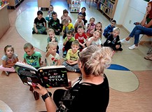 Na zdjęciu przedszkolaki siedzą na podłodze i słuchają Gosi czytającej 