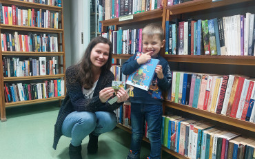 Bibliotekarka z chłopcem prezentują książkę, w tle regały z książkami