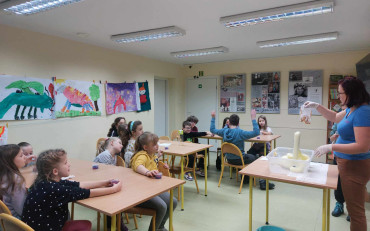 Dzieci siedą przy stolikach i obserwują doświadczenie prezentowane przez prowadzącą