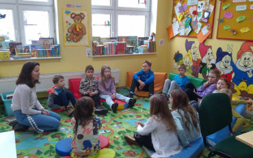 Dzieci siedzą na kolorowym dywanie i słuchają bibliotekarki opowiadającej historie