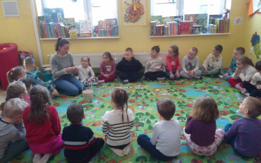 Przedszkolaki siedzą w kole na dywanie, a bibliotekarka czyta zagadkę