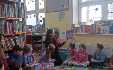 Dzieci siedzą na dywanie i słuchają opowiadania czytanego przez bibliotekarkę