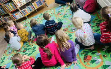 Dzieci siedzą na dywanie i słuchają opowiadania czytanego przez bibliotekarkę
