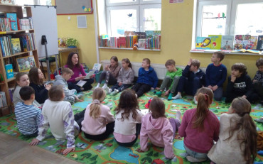 Dzieci siedzą na dywanie i słuchają opowiadania czytanego przez bibliotekarkę