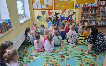 Dzieci siedzą na kolorowym dywanie i słuchają bibliotekarki, opowiadającej historie