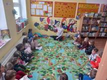 Dzieci siedzą w kole na dywanie, a Pani bibliotekarka czyta im książkę