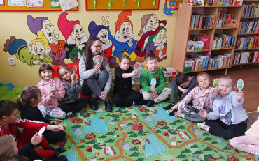 dzieci wraz z bibliotekarką siedzą w kole na dywanie i pokazują znalezione kolorowe kwiatki