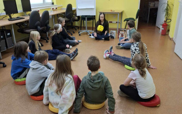Dzieci wraz z bibliotekarką siedzą w kole i rzucają kostką