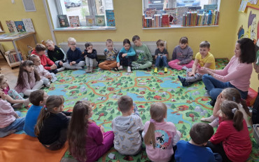 Dzieci wraz z bibliotekarką siedzą na kolorowym dywanie