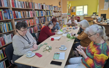 Panie siedzą przy stole, robią na drutach, rozmawiają.