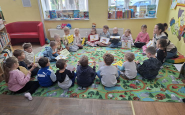 Dzieci wraz z bibliotekarką siedzą w kole na kolorowym dywanie