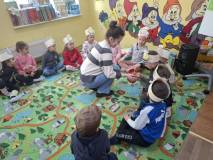 Bibliotekarka rozdaje dzieciom cukierki, dzieci w opaskach z pszczółką siedzą w kole na kolorowym dy