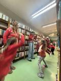 Dzieci wraz z paniami bibliotekarkami stoją między regałami z książkami i wykonują podskoki.