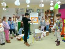 Dzieci stoją na środku sali  i obserwują chłopca siedzącego w kartonie. kolejny chłopiec rozplątuje 