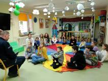 bibliotekarka wraz z dziećmi siedzi na kolorowej chuście. Dzieci patrzą na Pana Policjanta, który si