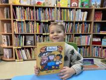 Chłopczyk siedzi na krześle, uśmiecha się do zdjęcia. w rękach trzyma książeczkę.