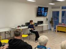 Zbliżenie na prowadzącą spotkanie. Młoda kobieta o brązowych włosach, ubrana w ciemny sweter i białą