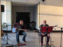 Dwóch mężczyzn siedzi na krzesłach w sali wystawowej podczas występu muzycznego. Mężczyzna po lewej 