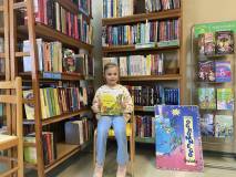 Dziewczynka siedzi na krześle, trzyma książkę w rekach. W tle są regały z książkami.