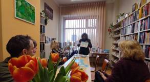 Uczestnicy spotkania przy stole w czytelni słuchają prelegentki, która pokazuje planszę edukacyjną. 